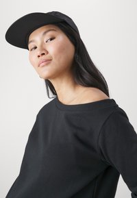 Črna majica s kapuco z off-shoulder oblikovanjem, z rebrastim izrezom in dolgimi rokavi. Oblečena z čepico, ki prikazuje sproščen kroj.