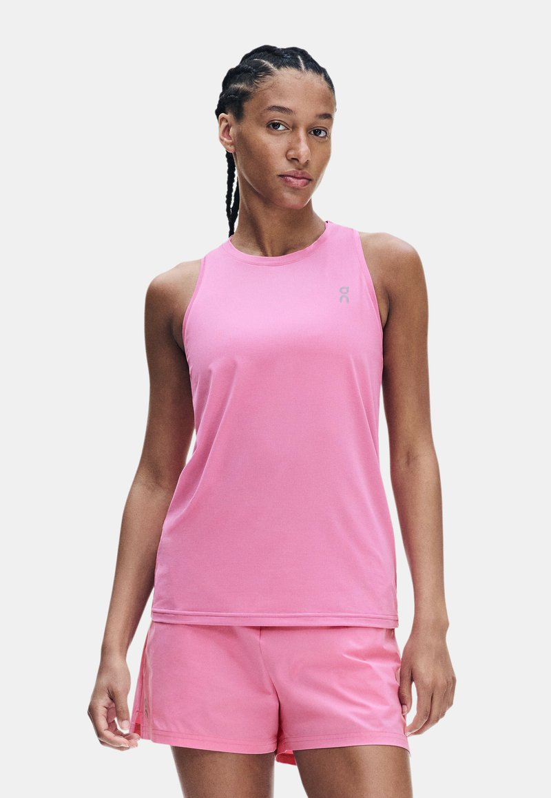Femme aux cheveux tressés portant un haut de sport rose sans manches et un short rose assorti, debout devant un fond uni.