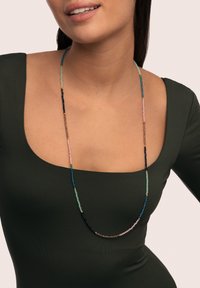 Collar largo de cuentas con cuentas azules, verdes, rosas y marrones con acentos dorados, colgando elegantemente sobre una blusa verde oscuro.