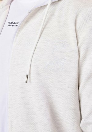 Sudadera con cremallera gris claro de tela texturizada, que presenta un logo bordado en blanco en el pecho. Incluye cordones con puntas plateadas.