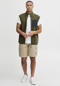 Oliven grønn fleecevest med en marineblå lomme, sammen med beige cargo-shorts. Modellen har på seg en hvit t-skjorte og hvite joggesko. Enkelt, funksjonelt design.