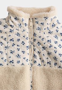 Giacca trapuntata floreale in crema con motivi blu, con colletto in pile e pannello inferiore. Chiusura con zip sul davanti, texture morbida.