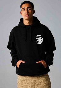Sudadera negra con bolsillo frontal, capucha con cordón y un diseño gráfico blanco en el lado izquierdo. El modelo lleva una prenda texturizada de color marrón.