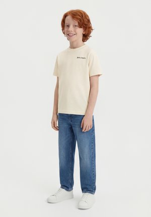 Vörös hajú fiú mosolyogva, krémszínű, rövid ujjú Palm Angels pólót, kék farmert és fehér tornacipőt viselve, egy egyszínű fehér háttér előtt áll.