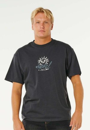 Blonde man met kort haar, gekleed in een zwart T-shirt met een zonafbeelding en gestileerde tekst op een witte achtergrond.