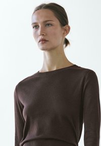 Jonge vrouw met lichte huid en bruin haar in een staart, gekleed in een donkerbruine top met lange mouwen, die iets schuin naar de zijkant kijkt tegen een effen achtergrond.