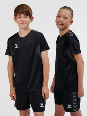 Twee glimlachende kinderen die zwarte Hummel sportshirts en korte broekjes dragen, staan tegen een effen lichte achtergrond.