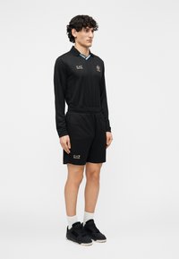 Črna športna oprava vključuje dolgotrajno majico s ovratnikom in logotipom ter črne kratke hlače. Detajli na ovratniku so v zlati in svetlo modri barvi.