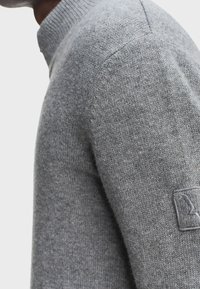 Close-up van een persoon die een grijze gebreide trui draagt met een geribbelde hoge kraag en een klein geborduurd patch op de bovenarm.