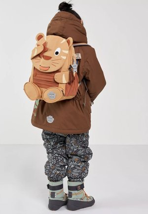 Kind trägt eine braune Jacke, gemusterte Hose, mintgrüne Stiefel und einen hellbraunen tierförmigen Rucksack, blickt mit dem Rücken zur Kamera vor einem einfarbigen Hintergrund.