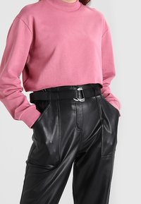 Persona con un sudadera corta rosa metida en pantalones de piel negra de cintura alta y un cinturón negro, con las manos en los bolsillos.