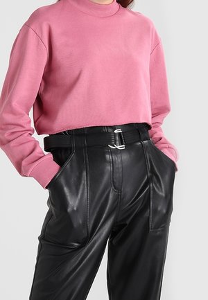 En person iklädd en rosa kort sweatshirt instoppad i högmidjade svarta läderbyxor med ett svart bälte, händerna i fickorna.