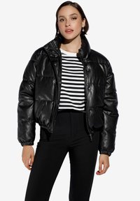 Chaqueta acolchada negra con un acabado brillante similar al cuero, cuello alto, cierre de botones y cremallera, llevada sobre una camiseta de rayas en blanco y negro.