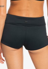 Svarte ribbede badeshorts med en tettsittende livlinje og en liten logoetikett. Materialet ser ut til å være elastisk og teksturert.