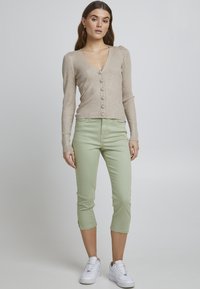 Cardigan beige à côtes avec décolleté en V et boutons, associé à un pantalon court vert clair avec fentes latérales, complété par des baskets blanches.