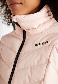 Světlá růžová prošívaná bunda s vysokým límcem, vybavená černým zipem a logem. Materiál má hladkou texturu s prošívanými detaily.