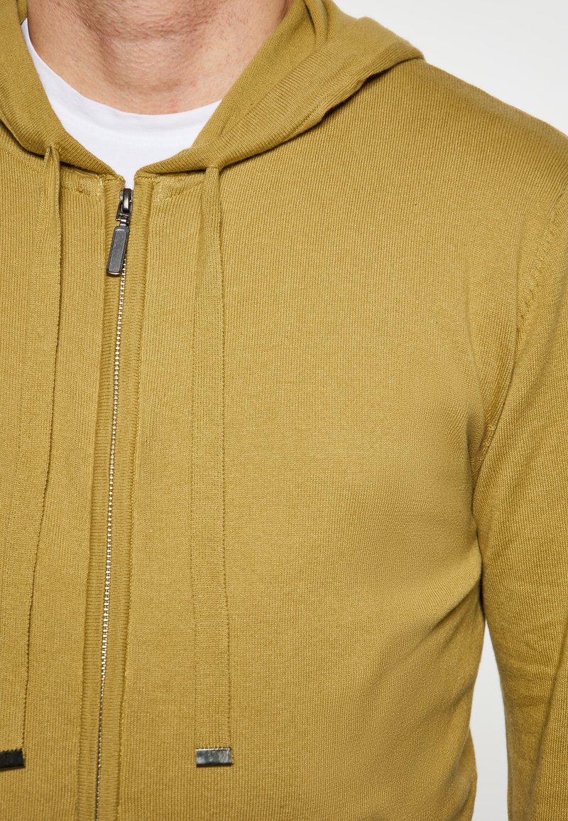Maglione con cappuccio aderente in tessuto a coste giallo senape. Presenta una chiusura frontale con cerniera e dettagli con lacci in metallo argentato.