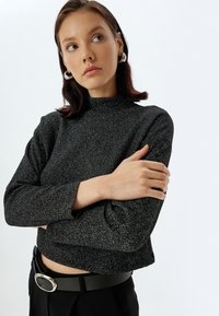 Schwarzes, glitzerndes Cropped Top mit hohem Kragen, langen Ärmeln und strukturierter Oberfläche. Kombiniert mit hochgeschnittenen schwarzen Hosen und einem Gürtel.