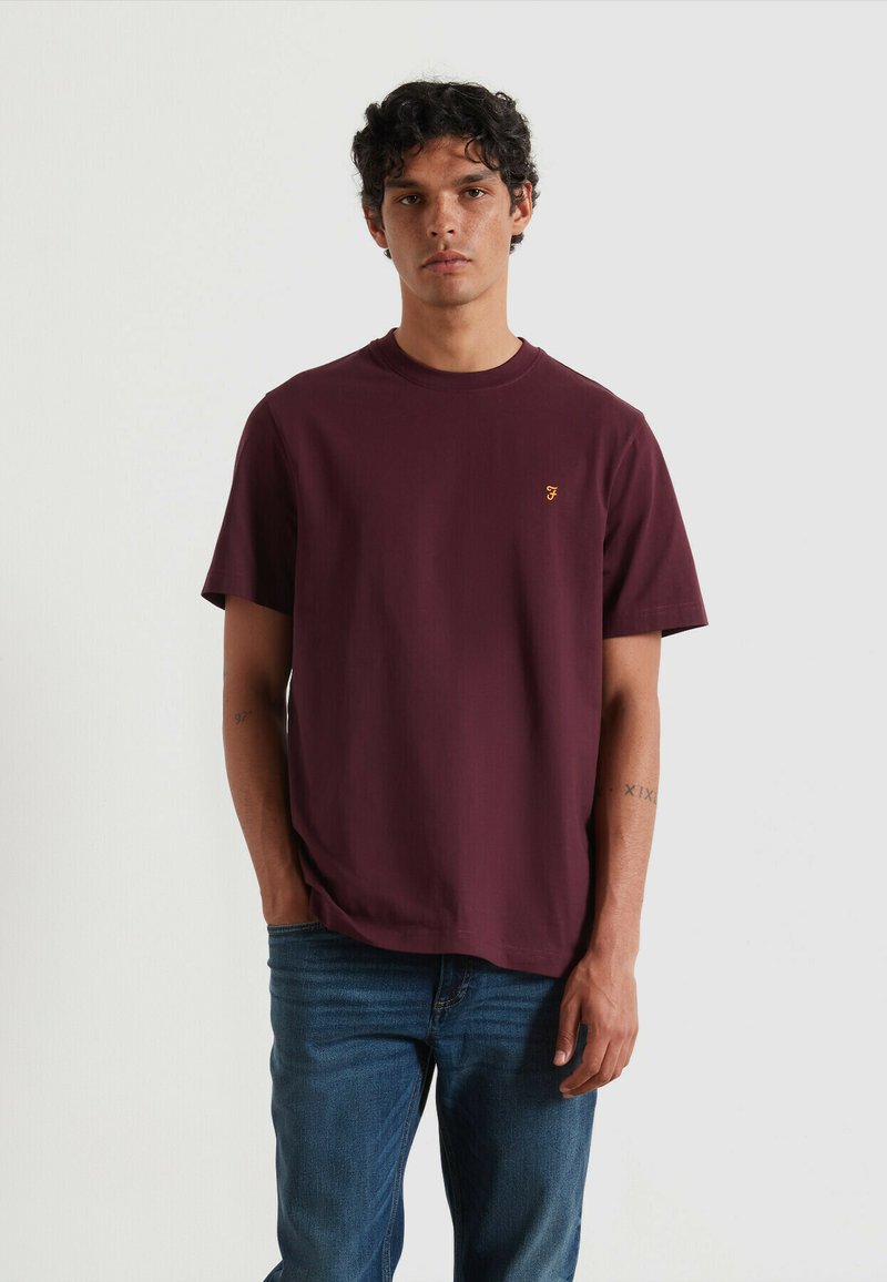 T-shirt di cotone color bordeaux con scollo rotondo e maniche corte, dotato di un piccolo logo giallo sul petto sinistro. Visibili jeans in denim.