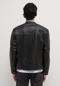 Uomo con capelli ricci scuri che indossa una giacca di pelle nera con spalle imbottite e jeans neri, rivolto verso uno sfondo chiaro e semplice.