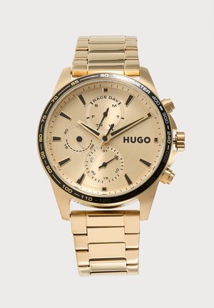 Montre-bracelet en or avec trois sous-cadrans, lunette tachymètre noire, et logo "HUGO", avec un bracelet métallique doré à maillons et deux boutons latéraux.