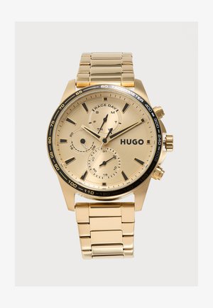 Zlaté náramkové hodinky s tromi subciferníkmi, čiernym tachymetrickým rámom a logom "HUGO", so zlatým kovovým remienkom a dvoma bočnými tlačidlami.