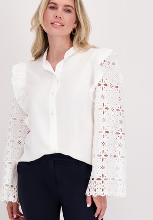 Witte blouse met rimpelkragen en gedetailleerde kant lange mouwen, met bloemen uitsparingen; gedragen met marineblauwe broek.