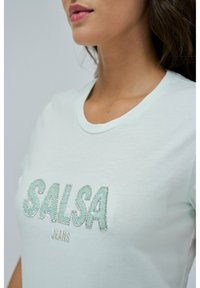 Světle mentolové tričko s krátkým rukávem a kulatým výstřihem. Na přední straně je nápis "SALSA" vytištěný ve třpytivých kamínkách a pod ním je vyšité "JEANS".
