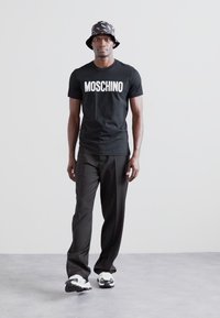 Svart Moschino T-skjorte med hvit tekst, kombinert med løse svarte bukser og mønstret svart bøttehatt. Sporty hvite og svarte sneakers.