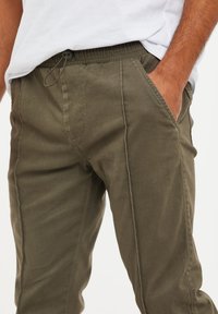 Pantalons verts olive fuselés avec une taille élastique, poches latérales et un cordon de serrage pour l'ajustement. Tissu lisse avec une texture minimale.