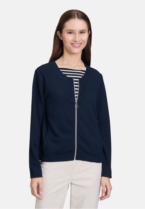 Veste zippée navy avec texture matelassée, col rond, manches longues et ourlet côtelé. Portée par-dessus un haut rayé et un pantalon clair.
