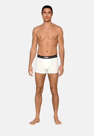 Mann mit langen dunklen Haaren steht barfuß in weißen Boxershorts mit schwarzem Bund und der Aufschrift "JOOP!" vor einem einfarbigen Hintergrund.