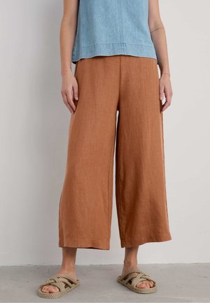 Persona con pantalones cortos marrones de pierna ancha, camiseta sin mangas azul claro y sandalias beige de cuerda, de pie frente a una pared lisa y clara.