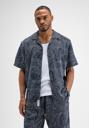 Mann trägt dunkelblau gemustertes kurzärmeliges Hemd und passende Hose über einem weißen Tanktop, steht mit einer Hand in der Tasche.
