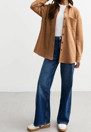 Person trägt ein beige Button-up-Hemd, weißes Top, weite blaue Jeans und weiße Sneaker und steht vor einem schlichten hellen Hintergrund.