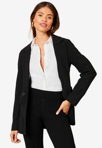 Černý přiléhavý blazer s klopami, zapínáním na jeden knoflík a dlouhými rukávy, pod kterým je bílé button-up tričko.