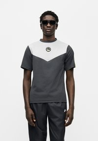 Μαύρο και γκρι κοντομάνικο t-shirt με σχέδιο με σχήμα βέλους, στρογγυλή λαιμόκοψη και λογότυπο. Φορεμένο με σκούρο παντελόνι και γυαλιά ηλίου.