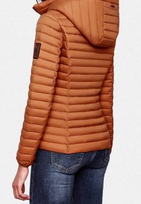 Prošívaná oranžovo-hnědá péřová bunda s přiléhavým střihem a kapucí. Má dlouhé rukávy a texturovanou tkaninu. Nositelná přes modré džíny.