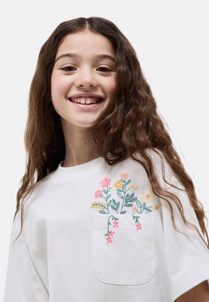Fille souriante aux longs cheveux bruns portant une chemise blanche avec une poche brodée de fleurs colorées.