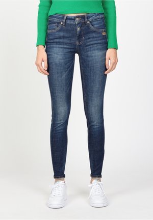 Femme portant un jean skinny bleu foncé, des baskets blanches et un haut vert à manches longues, debout devant un fond blanc uni.