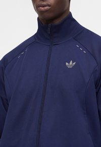 Άτομο που φοράει μπλε σκούρο Adidas αθλητικό τζάκετ με φερμουάρ, ψηλό γιακά, ανακλαστικό λογότυπο στο αριστερό στήθος και ανακλαστικές γραμμές στους ώμους.