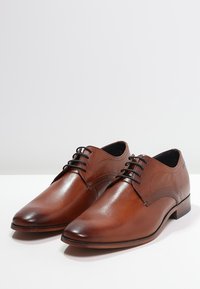 Paire de chaussures élégantes en cuir marron poli avec des lacets noirs sur une surface blanche, sur un fond clair uni.