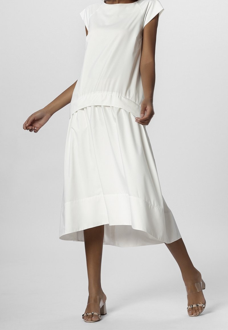 Femme portant une robe blanche fluide sans manches avec un ourlet asymétrique et des sandales à talons blocs avec brides transparentes sur un fond uni.