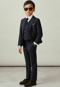 Completo blu navy a tre pezzi con gilet, camicia bianca e cravatta blu navy. Mocassini in suede marrone. Occhiali da sole neri e fazzoletto da taschino con motivi.