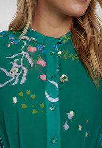 Zöld, gombos blúz, álló gallérral, rózsaszín, sárga és arany díszítéssel ellátott virágmintákkal. Áttetsző, sima anyag.