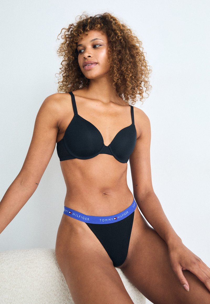 Zwarte gewatteerde bh met verstelbare straps en een gladde textuur, gecombineerd met een zwarte string met een blauwe "TOMMY HILFIGER" logo tailleband.