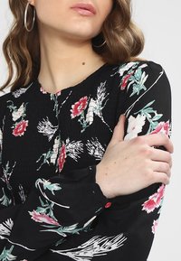 Svart blommig blus med långa ärmar som har en volangstruktur. Designen inkluderar rosa och gröna blommönster på en mörk bakgrund.