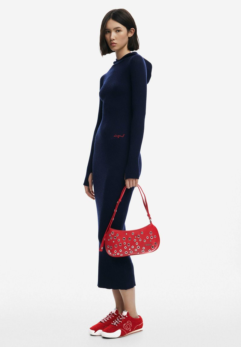 Rochie din material filetat navy cu glugă, detalii brodate roșii, asortată cu o geantă roșie cu accente cu găuri și pantofi sport roșii cu tălpi albe.