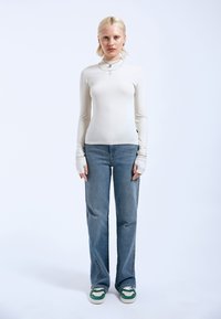 White long-sleeve top with thumbholes, paired with light blue flared jeans. Footwear features green and white accents. Simple design elements.