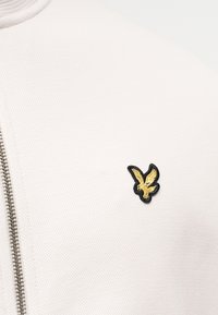 Λευκό υφασμάτινο μπουφάν με ασημένιο φερμουάρ και μικρό κίτρινο πουλί κεντημένο στο στήθος.